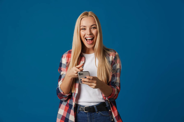 Blonde happy white woman holding and pointing finger at mobile phone isolated over blue background - Fotó, kép