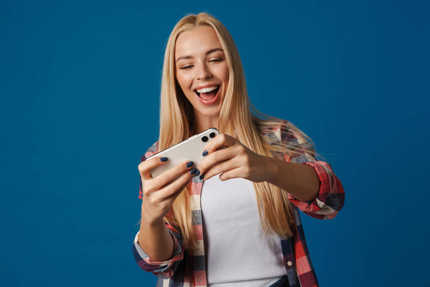 Happy beautiful blonde girl playing online game on smartphone isolated over blue background - Photo, image