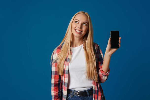 Vidám szőke lány mosolyog és mutatja okostelefon elszigetelt felett kék háttér - Fotó, kép