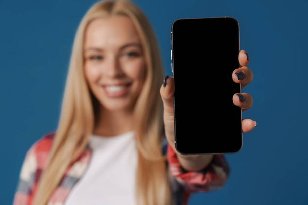 Bionda felice donna bianca sorridente mentre mostra il telefono cellulare isolato su sfondo blu - Foto, immagini