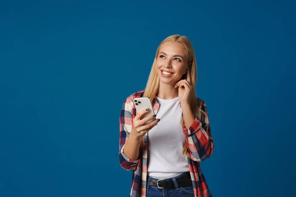 Blonde happy white woman using mobile phone and wireless earphone isolated over blue background - Фото, изображение