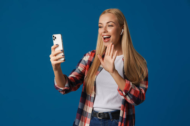 Blonde happy woman gesturing while using mobile phone and earphone isolated over blue background - Fotó, kép