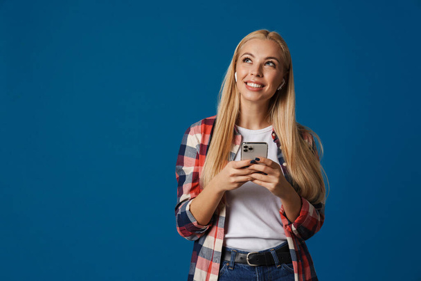 Cheerful beautiful blonde girl smiling and using smartphone isolated over blue background - Fotó, kép