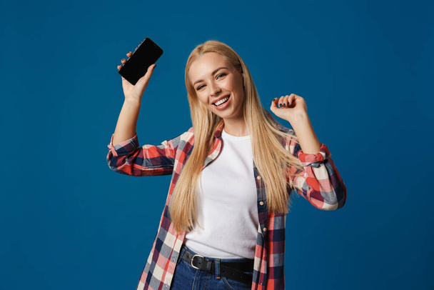 Cheerful beautiful blonde girl smiling while dancing with smartphone isolated over blue background - Φωτογραφία, εικόνα