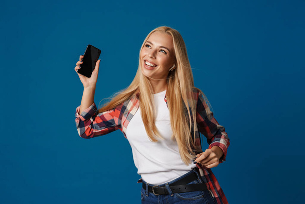Cheerful beautiful blonde girl smiling while dancing with smartphone isolated over blue background - Photo, Image