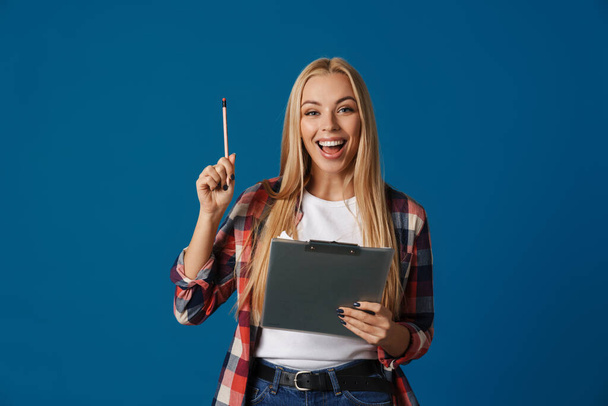 Blonde excited woman smiling while posing with clipboard and pencil isolated over blue background - Foto, imagen