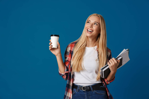 Blonde glückliche Frau lächelt, während sie mit Kaffee und Notizbuch vor blauem Hintergrund posiert - Foto, Bild