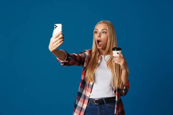 Shocked girl taking selfie on smartphone while drinking coffee isolated over blue background - Foto, imagen