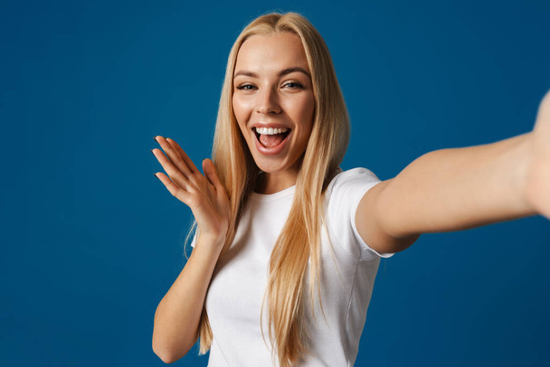 Blonde young happy woman smiling while taking selfie photo isolated over blue background - Fotó, kép