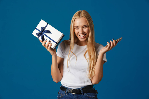 Happy beautiful girl posing with mobile phone and gift box isolated over blue background - Zdjęcie, obraz