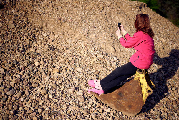 Photo of a caucasian woman in a jacket outdoors on a background of sand looking at the phone while sitting - Fotó, kép