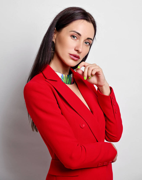 Portrait of businesswoman with permanent makeup in red jacket and modern jewelry standing sideways, looking at camera - Fotó, kép