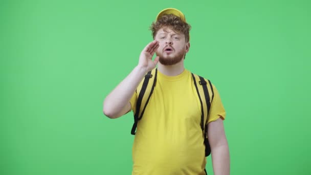 Portrait of young men screaming calling someone. Redhead male in yellow delivery courier uniform with thermo bag posing on green screen in studio. Close up. Slow motion ready 59.97fps. - Video