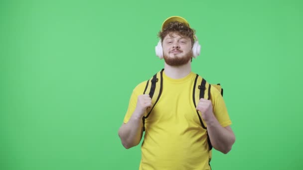 Portrait of young men enjoying music in big white headphones. Redhead male in yellow delivery courier uniform with thermo bag posing on green screen in studio. Close up. Slow motion ready 59.97fps. - Materiał filmowy, wideo