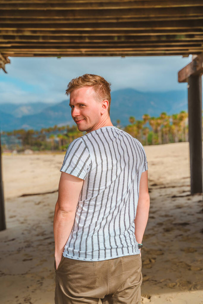 A young man poses under a pier on the beach against the backdrop of palm trees in Santa Barbara, California, USA - Valokuva, kuva