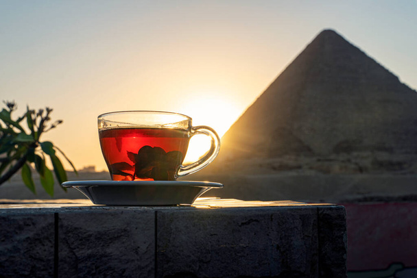 За мятным чаем в кафе, размышляя о впечатляющих трех пирамидах Гизы. Прозрачная стеклянная кружка с черным чаем и мятой на фоне египетских пирамид. - Фото, изображение