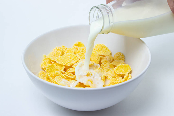La leche se vierte en un plato con copos de maíz sobre un fondo blanco. Desayuno saludable y sabroso. - Foto, Imagen