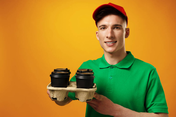 Young delivery man holding coffee cups against yellow background - Valokuva, kuva