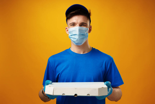 Young delivery man in face mask and gloves holding food delivery parcel against yellow background - Foto, Bild