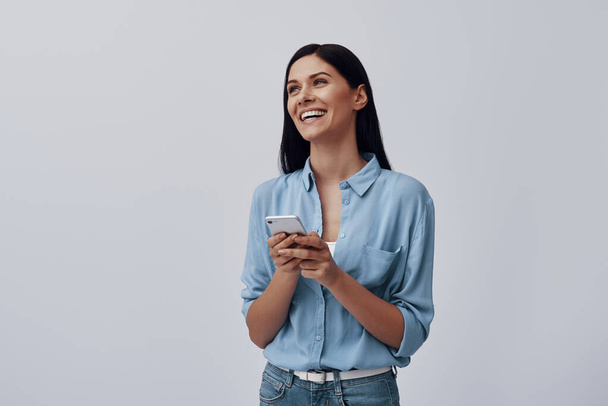 Attractive young woman using smart phone and smiling while standing against grey background - Φωτογραφία, εικόνα
