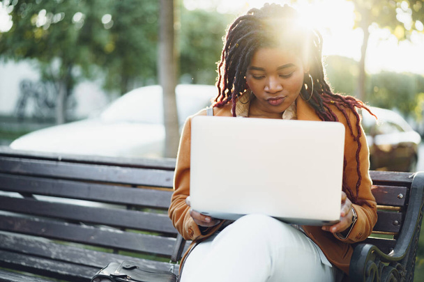 African american woman freelancer sitting on a bench in a street and using laptop - Foto, Imagen