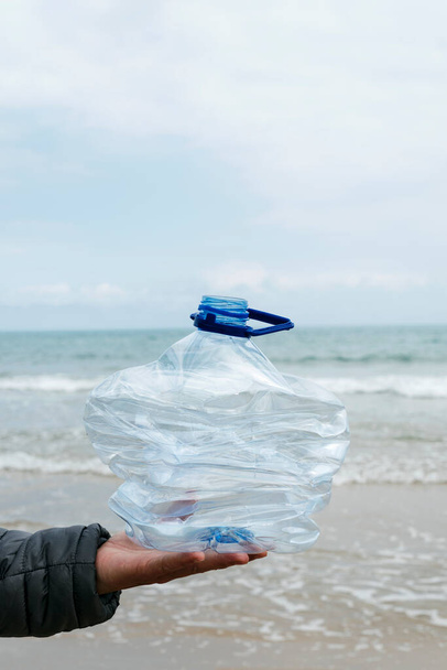 gros plan d'un jeune homme avec une bouteille en plastique usagée broyée à la main, fraîchement recueillie dans le sable d'une plage, comme une action pour nettoyer l'environnement naturel - Photo, image
