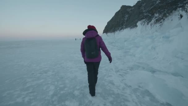 Une jeune fille marche le long du lac Baïkal gelé au-delà des blocs de glace et des montagnes rocheuses sur le rivage. Vue de derrière. Mouvement lent - Séquence, vidéo