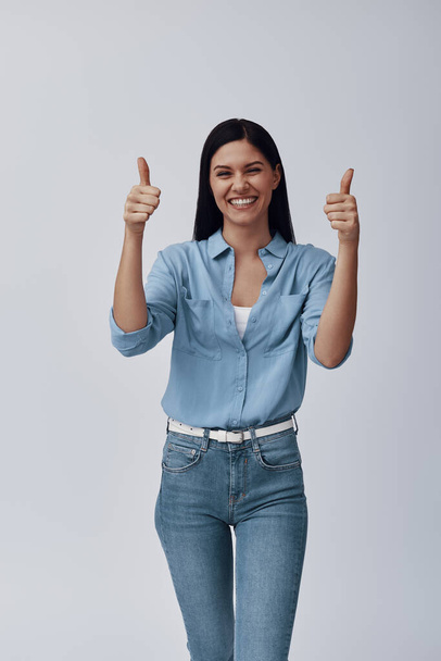 Attractive young woman looking at camera and keeping thumbs up while standing against grey background - Fotoğraf, Görsel