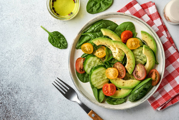 Avocado, red, yellow, black cherry tomato, spinach and cucumber fresh salad with spices pepper and olive oil in grey bowl on grey slate, stone or concrete background. Healthy food concept. Top view. - Foto, Bild