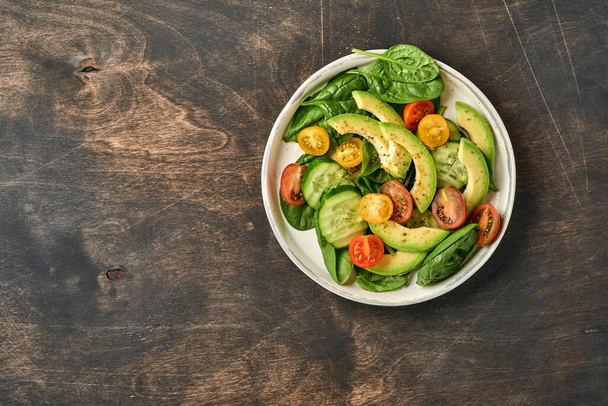 Avocado, red, yellow, black cherry tomato, spinach and cucumber fresh salad with spices pepper and olive oil in grey bowl old wooden table background. Healthy food concept. Top view. - Фото, зображення