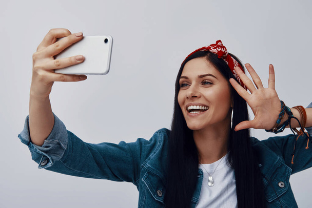 Funky young woman in bandana making selfie and smiling while standing against grey background - Photo, Image