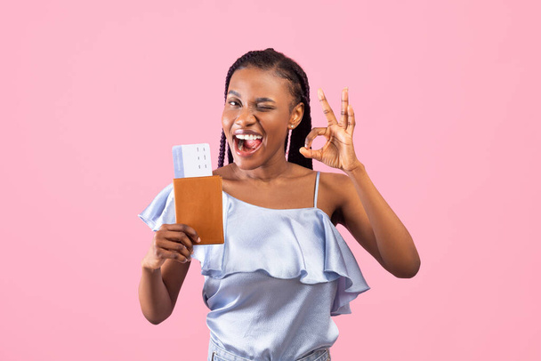 Positive black woman holding passport and plane tickets, showing okay gesture and winking over pink background - Zdjęcie, obraz