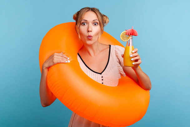 Blonde adorable woman with astonished facial expression looking directly at camera and combines lips a tubule, holding rubber ring and cocktail. Indoor studio shot isolated on blue background. - 写真・画像