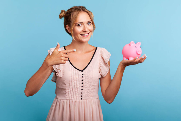 Smiling positive female with hair buns in summer dress holding and pointing at piggy bank with finger, suggesting you to put coins inside, saving money. Indoor studio shot isolated on blue background. - Foto, afbeelding
