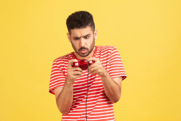 Addicted nervous bearded man in red striped t-shirt grimacing holding joystick, playing video games online, virtual competition. Indoor studio shot isolated on yellow background - Photo, image