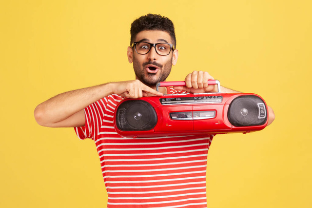 Excited happy bearded man in striped t-shirt and fashionable eyeglasses holding in hand red tape recorder, pushing play button. Indoor studio shot isolated on yellow background - Фото, зображення