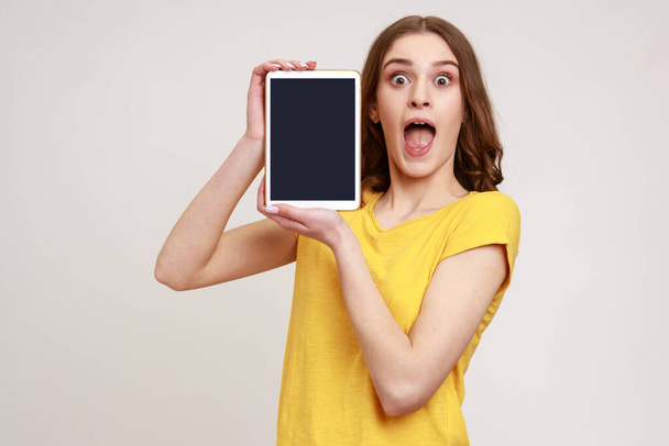 Young excited woman wearing casual yellow T-shirt, holding digital tablet in hands, showing blank device screen to camera, expressing astonishment. Indoor studio shot isolated on gray background. - Фото, изображение