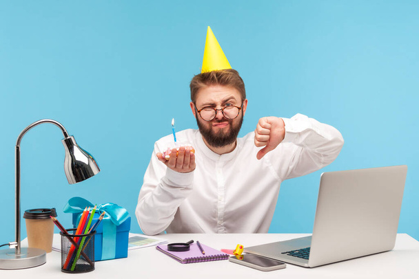 Rude unhappy man with beard in eyeglasses and cone hat showing thumbs down holding cake with candle, dissatisfied with anniversary celebration at workplace. Indoor studio shot isolated on blue background - Foto, immagini