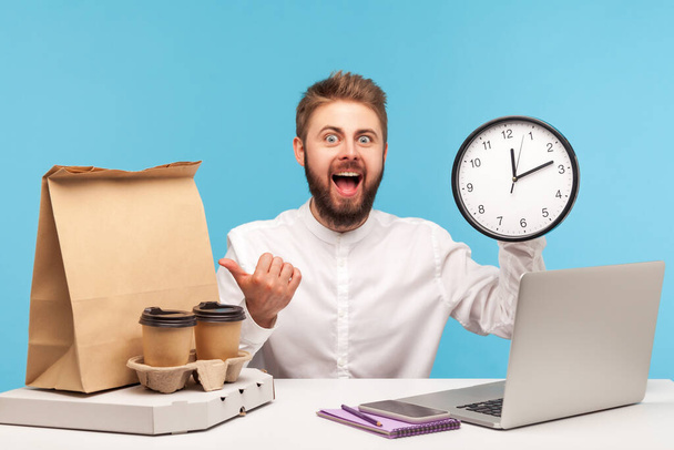 Excited positive bearded man office worker holding big wall clock pointing finger at paper bag, coffee cups and pizza box, satisfied with fast delivery. Indoor studio shot isolated on blue background - Fotoğraf, Görsel