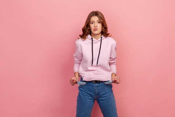 Portrait of upset poor student girl in hoodie and jeans turning out empty pockets, showing no money gesture, worried about debts. Indoor studio shot isolated on pink background - Photo, Image