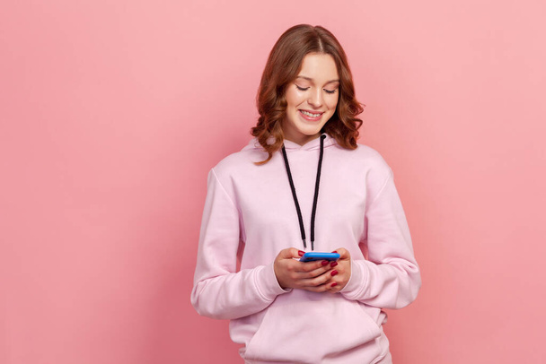 Portrait of cheerful brunette teenage female in casual hoodie sincerely smiling while using smartphone, messaging, entertainment app. Indoor studio shot isolated on pink background - Photo, Image