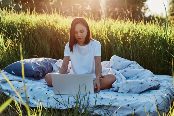 Outdoor shot of beautiful brunette female freelancer working with lap top in green meadow on soft bed, looking at lap top screen with concentrated expression. - Fotoğraf, Görsel