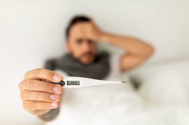 Fever. Ill man showing thermometer with high temperature result to camera, lying on bed. Selective focus - Фото, изображение