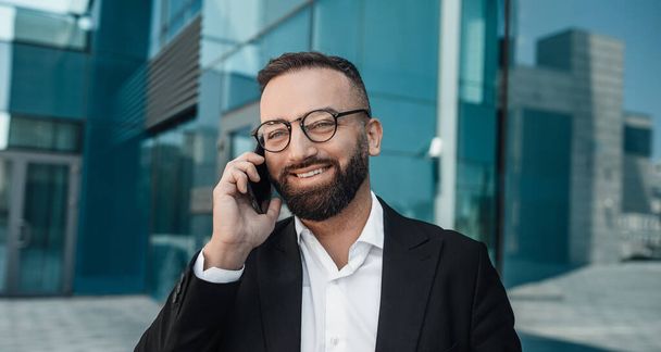 Business call. Confident bearded businessman chatting on cellphone standing outdoor in city - Photo, image