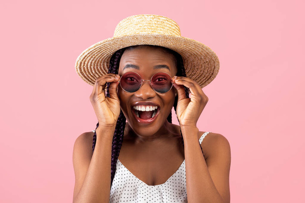 Femme afro-américaine millénaire excitée en tenue d'été touchant ses lunettes de soleil, criant WOW sur fond rose - Photo, image