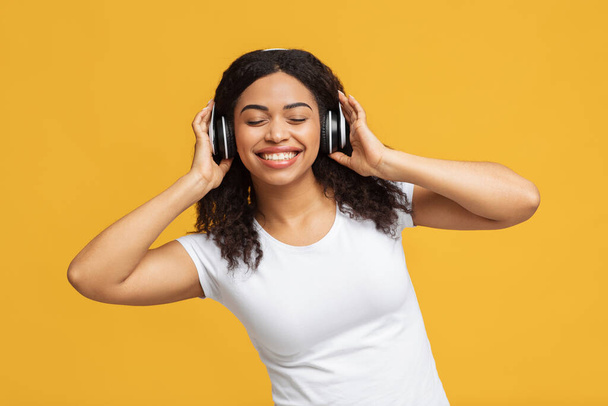 I like this tune. Positive black woman enjoying music with closed eyes, touching wireless headphones and dancing - Фото, изображение