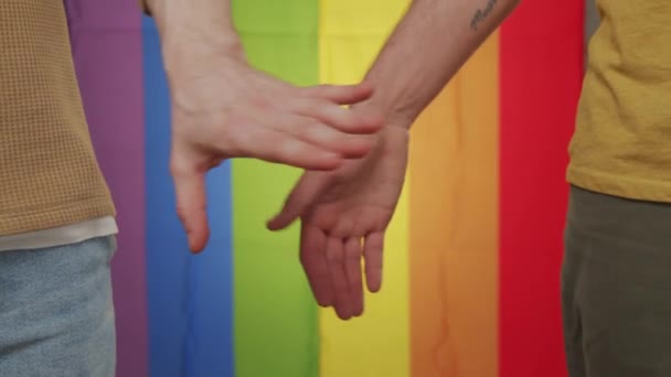 Κοντινό πλάνο αγνώριστου γκέι ζευγαριού που κρατιέται χέρι-χέρι πριν τη σημαία του ουράνιου τόξου - Πλάνα, βίντεο
