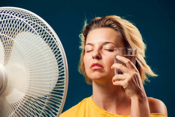 Ein Porträt einer Frau vor einem Ventilator mit einem Glas Wasser, das unter Hitze leidet. Konzept für heißes Wetter - Foto, Bild