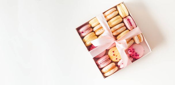 Flache Lagen gelber und pinkfarbener Makronen auf weißem Hintergrund mit freier Textfläche. Makroaufnahme von süßen französischen Dessertkeksen, die zusammen in der Geschenkbox arrangiert sind und mit einer Schleife gebunden sind. - Foto, Bild