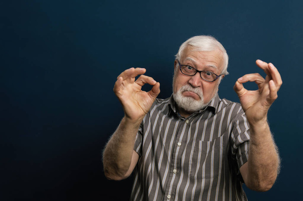 Carismático hombre de pelo gris en gafas muestra bien con ambas manos, emoción en la montura, retrato de media longitud, fondo azul - Foto, imagen